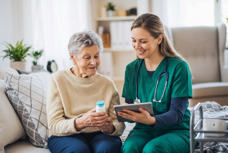 A nurse in green scrubs shows a tablet to an elderly woman holding a medicine bottle. They sit together on a sofa in a well-lit room, smiling and engaged in conversation.