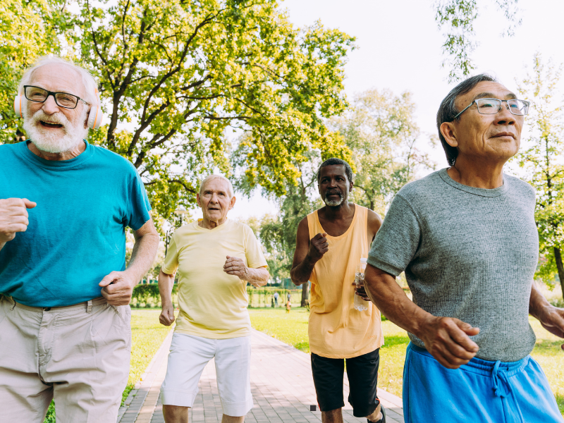 Four older men are jogging together outdoors in a park on a sunny day, surrounded by green trees and grass. They appear happy and active, wearing casual workout clothing.