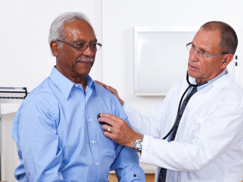 A doctor uses a stethoscope to listen to the chest of an older man wearing a blue shirt during a medical examination in a clinic.