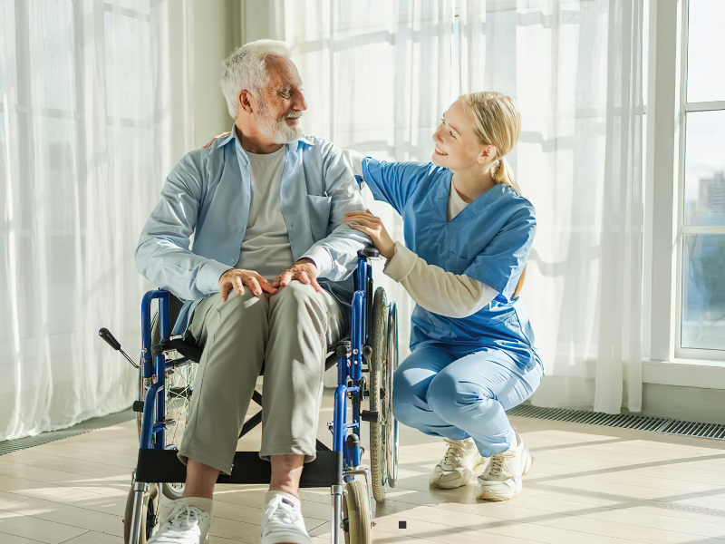 A smiling elderly man in a wheelchair sits beside a cheerful young nurse in blue scrubs, who kneels next to him with a hand on his shoulder. They are in a bright room with large windows and sheer curtains.