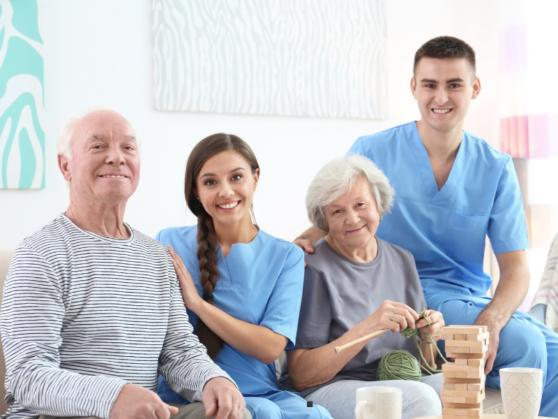 Two elderly people sit with two young caregivers in blue scrubs. The elderly woman is knitting while the elderly man smiles. Everyone looks happy and relaxed, with a Jenga game and mugs on the table in front of them.