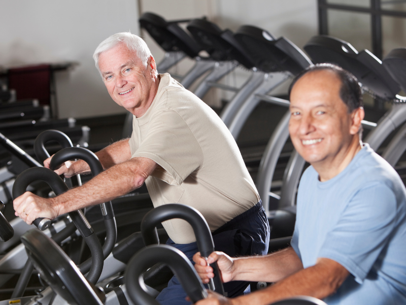 Two older men smiling while exercising on stationary bikes in a gym. They are wearing casual workout clothes and appear to be enjoying their fitness routine. Several more exercise machines are visible in the background.