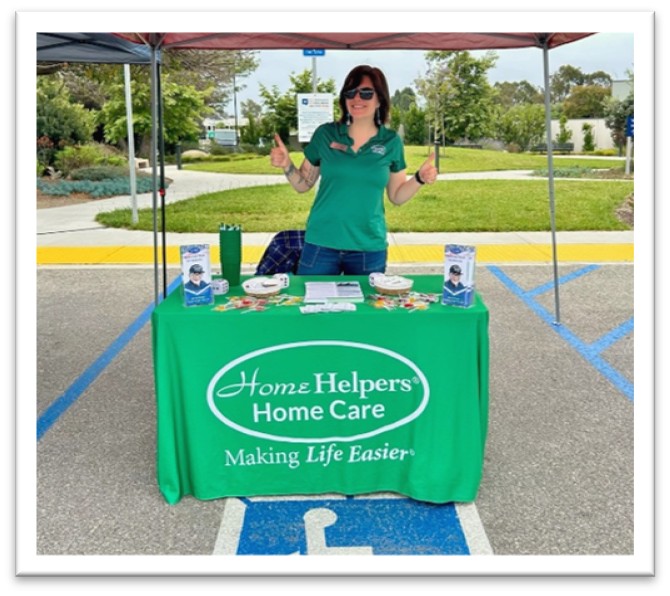 A woman in sunglasses and a green shirt stands behind a table covered with a green Home Helpers Home Care tablecloth at an outdoor event, giving two thumbs up. Pamphlets and giveaways are arranged on the table.