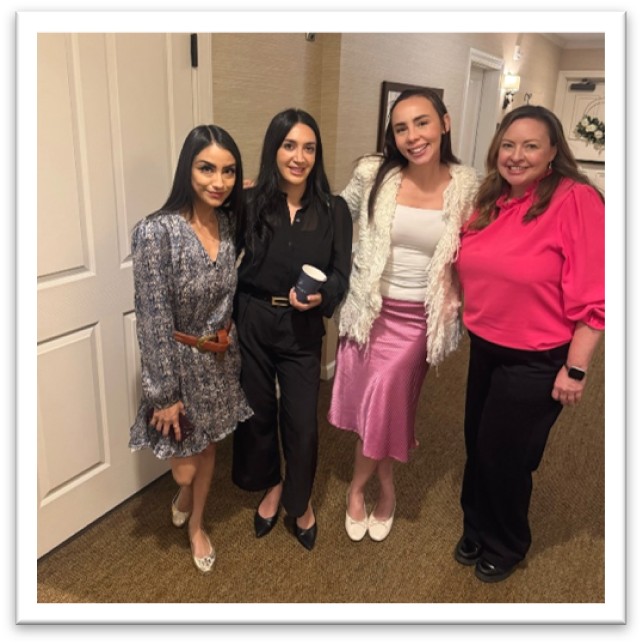 Four women stand together indoors, smiling at the camera. They are dressed in stylish outfits: patterned dress, black suit, white jacket with pink skirt, and a bright pink blouse. The background shows a hallway with beige walls.