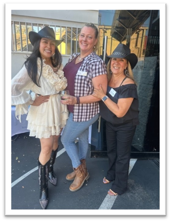 Three women wearing cowboy hats smile and pose together outdoors. One wears a white ruffled dress and boots, another wears jeans, a plaid shirt, and boots, and the third wears black pants and a black top.
