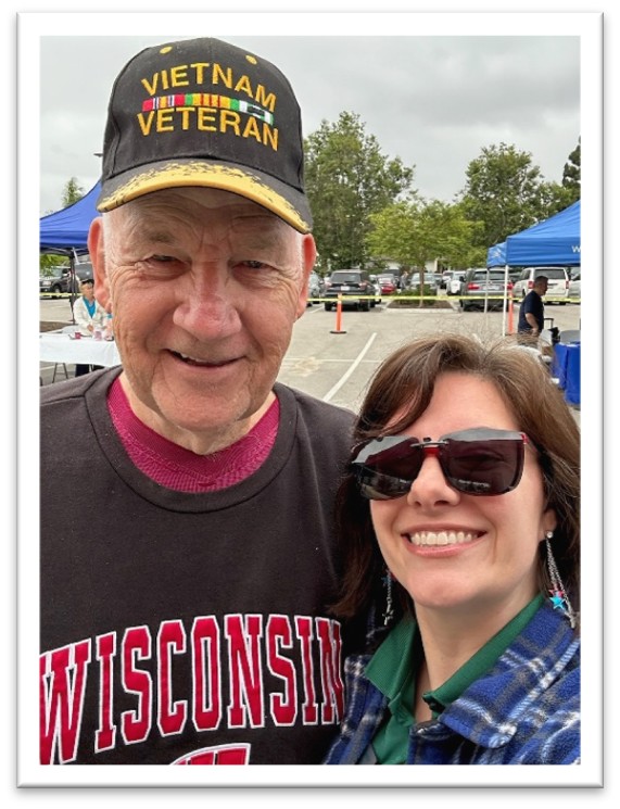 An older man wearing a Vietnam Veteran hat and a Wisconsin sweatshirt smiles next to a woman in sunglasses. They are outdoors, with trees, cars, and blue tents in the background.