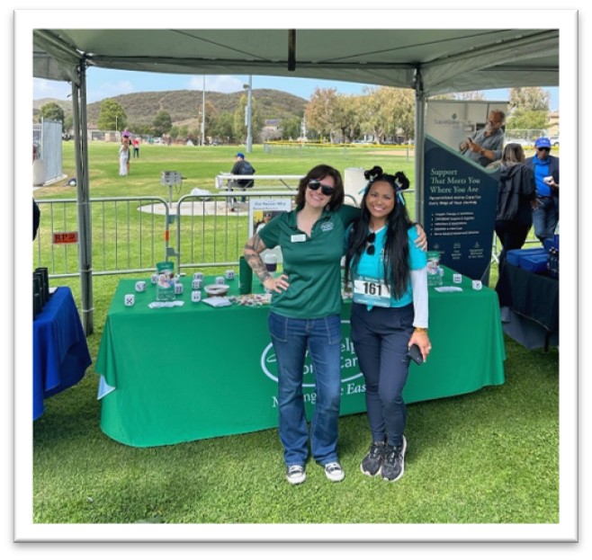 Two women stand smiling under a tent at an outdoor event, with a green table covered in promotional items and banners behind them. Grass, people, and hills are visible in the background.