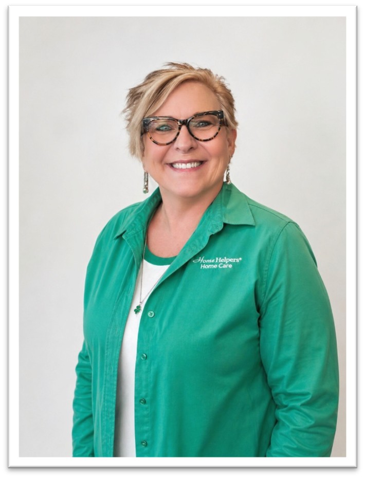 A smiling woman with short blonde hair, wearing glasses and a green Home Helpers Home Care shirt, stands against a plain light background.