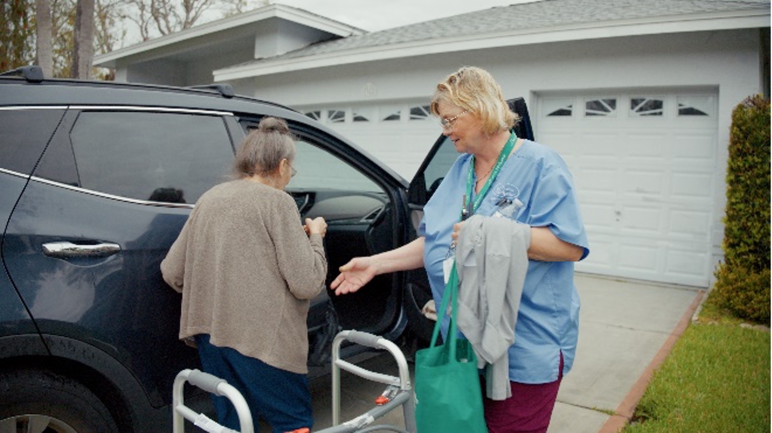 A caregiver in scrubs helps an elderly woman using a walker into a car parked in front of a suburban house with a double garage.