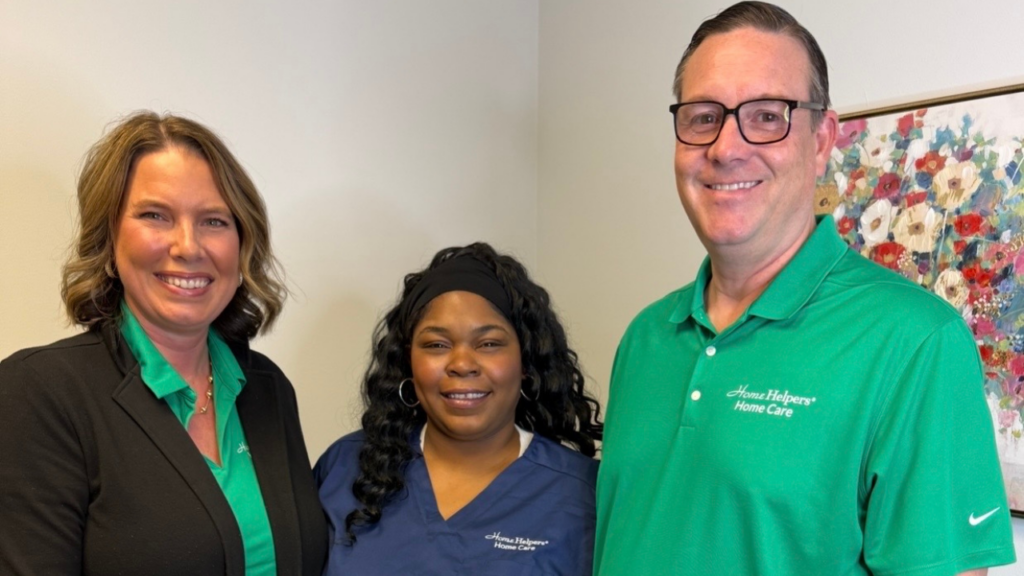 Three people stand together smiling. Two wear green shirts with Home Helpers Home Care logos, and one wears a navy blue scrub top with the same logo. A colorful floral painting hangs on the wall behind them.