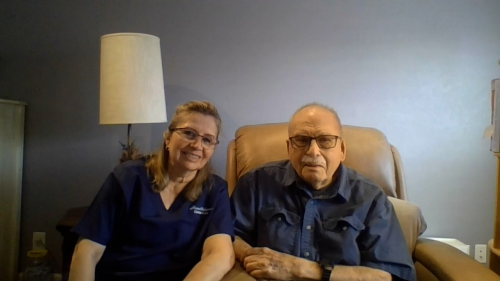 A woman and an older man sit together on a tan chair in a room with gray walls. The woman is smiling and wearing glasses and a navy top; the man is wearing glasses and a dark shirt. A lamp is visible in the background.