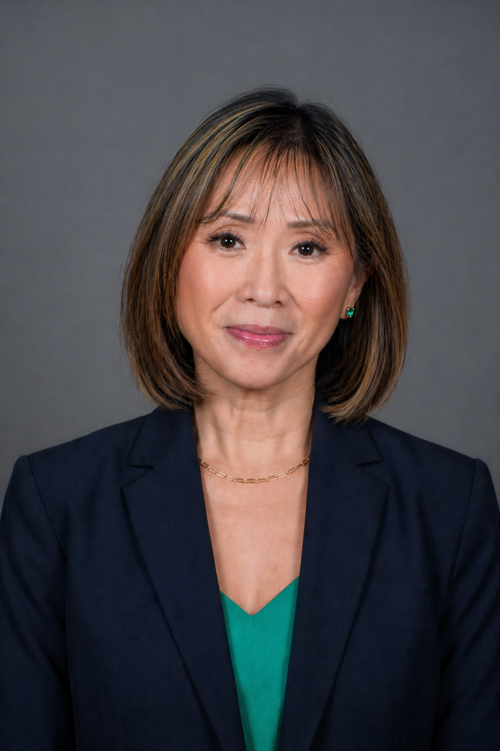 A woman with shoulder-length brown hair wearing a navy blazer and green blouse poses against a plain gray background, looking at the camera with a slight smile.
