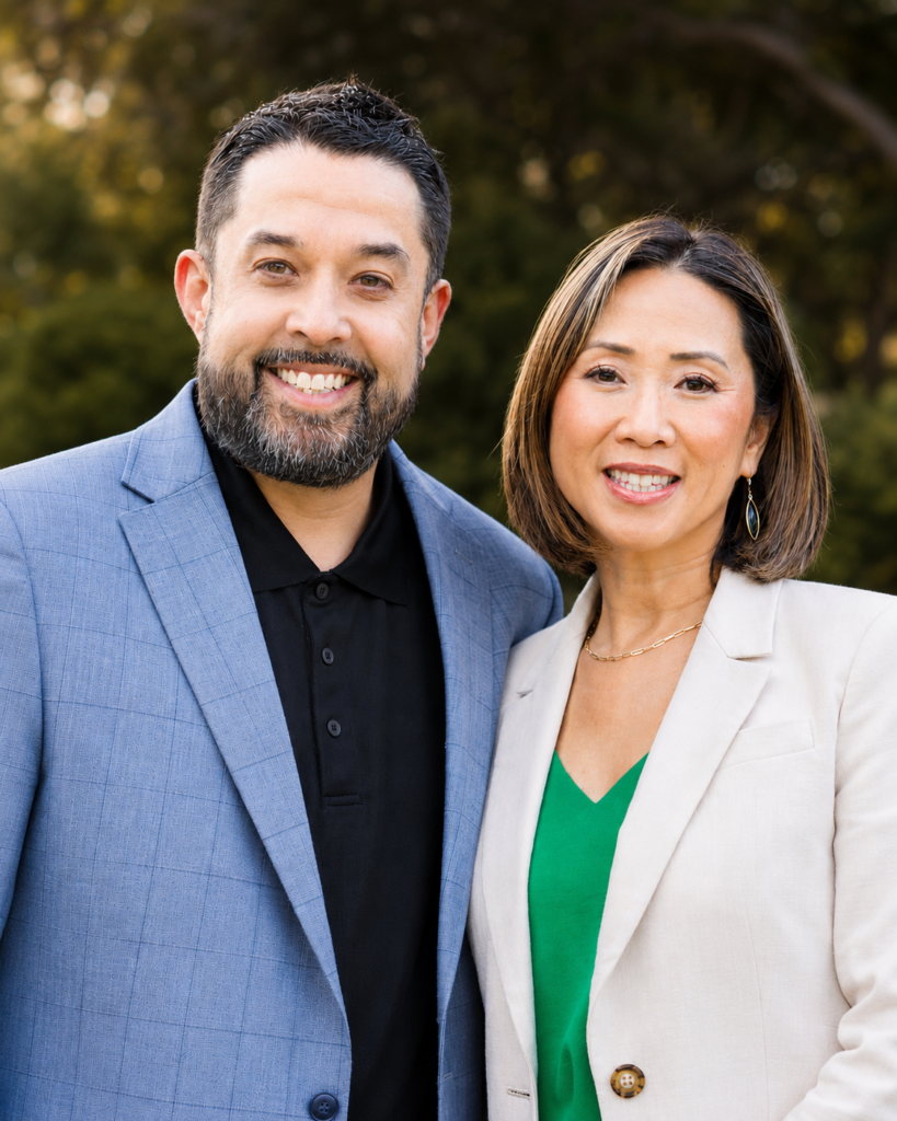 A man and woman stand side by side outdoors, both smiling at the camera. They wear blazers, with the man in a blue jacket and the woman in a light jacket over a green top. Green trees are blurred in the background.
