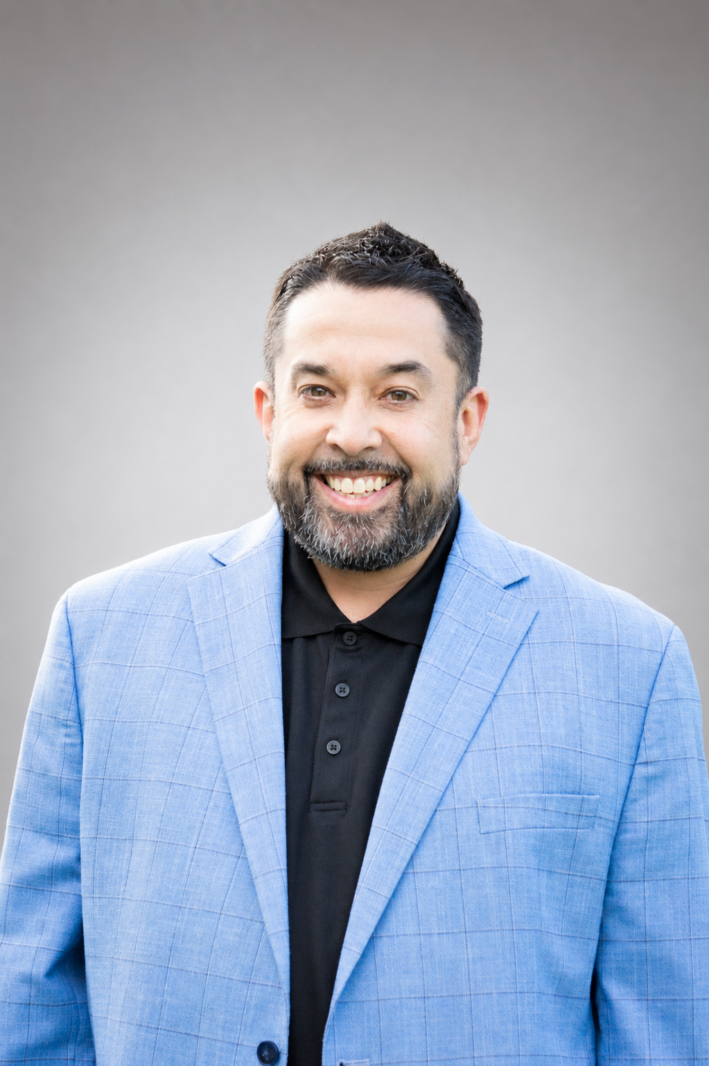 Smiling man with short dark hair and beard, wearing a light blue plaid blazer over a black collared shirt, standing in front of a plain gray background.