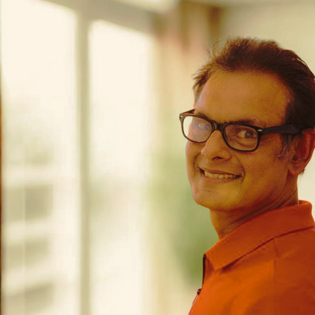 A man wearing glasses and an orange shirt smiles while looking over his shoulder in a brightly lit indoor setting.
