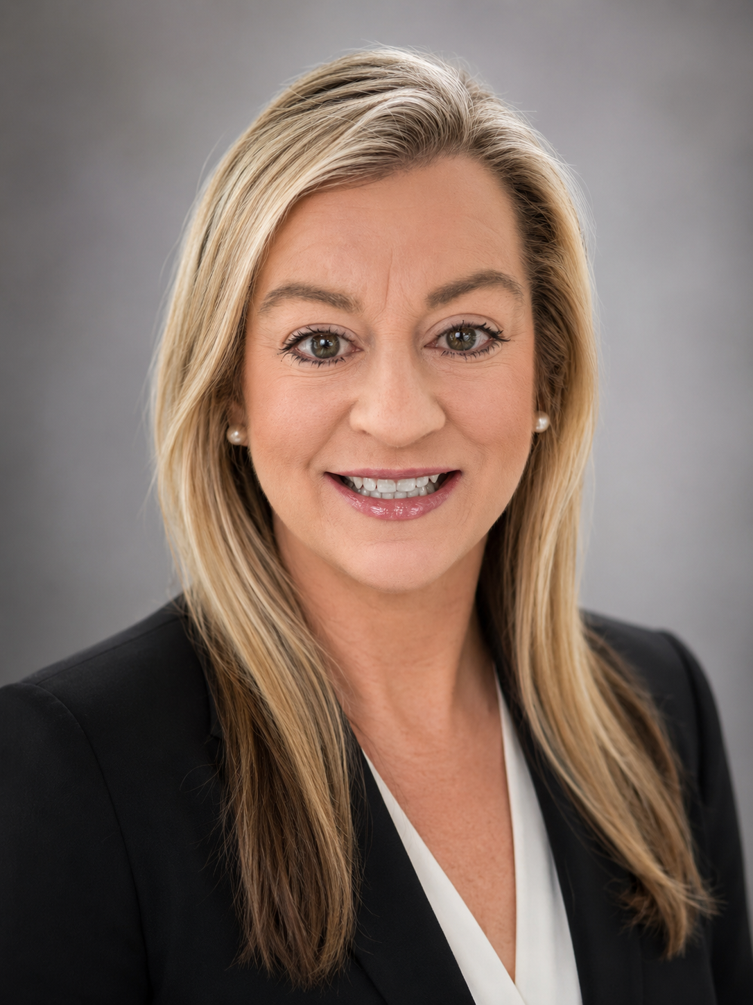 Professional woman with long blonde hair, wearing a black blazer and white blouse, smiling at the camera against a neutral gray background.