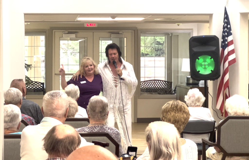A man dressed as Elvis Presley performs with a woman beside him in front of an audience of seniors in a bright room with an American flag and green stage light.