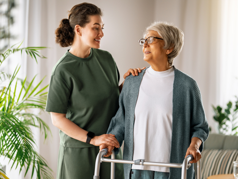 A young woman supports and smiles at an older woman using a walker indoors. They appear happy and are surrounded by plants and soft natural light.