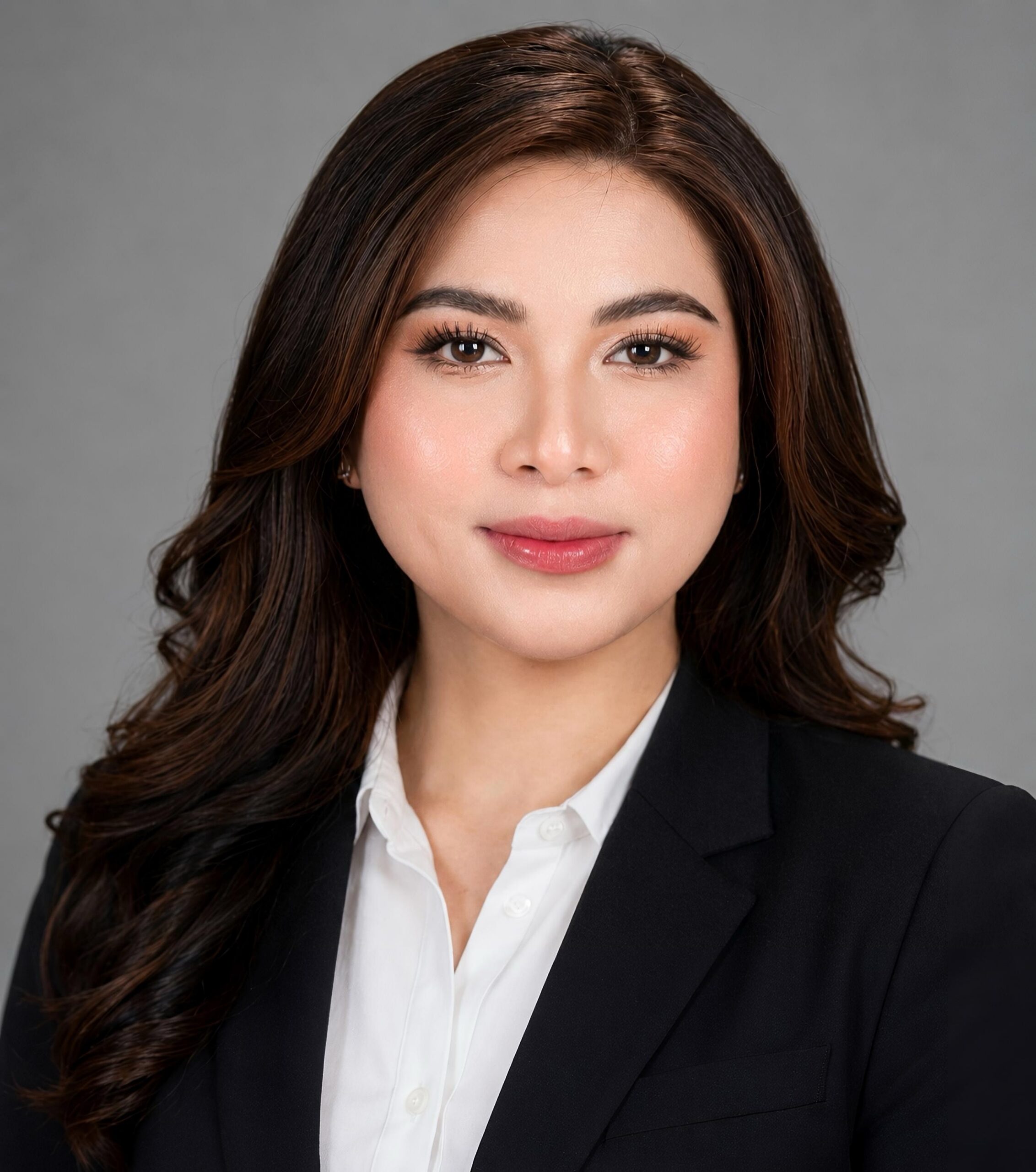 Professional portrait of a woman with long, wavy dark hair wearing a black blazer and white shirt, smiling softly against a plain gray background.