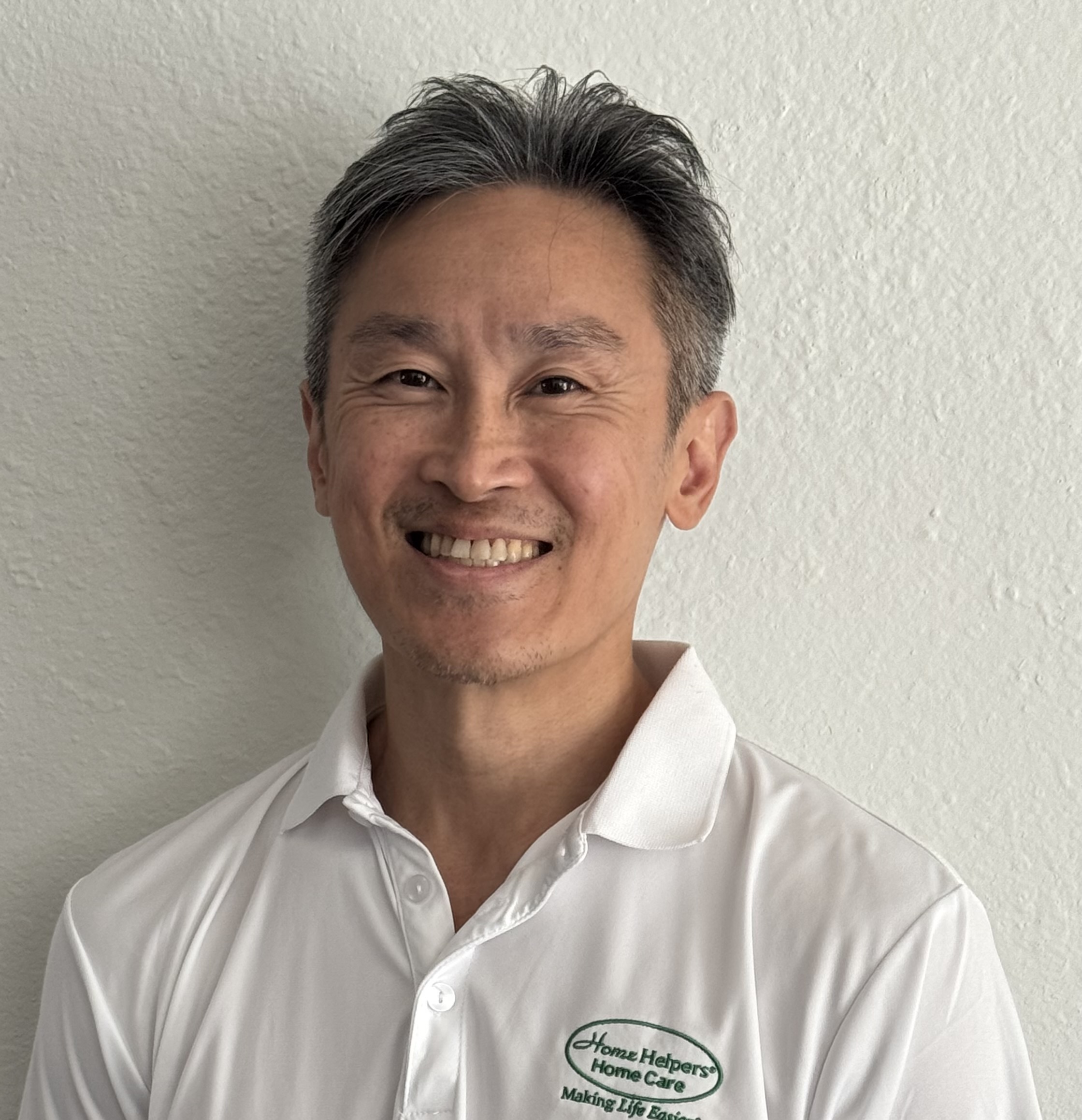 Smiling man with short gray hair wearing a white Home Helpers Home Care polo shirt stands in front of a light-colored wall.