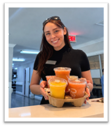 A woman with long brown hair, wearing black clothes and sunglasses on her head, smiles while holding a drink carrier with several colorful juice cups in a bright indoor setting.