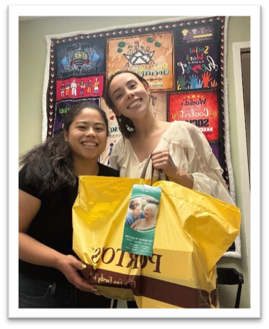 Two women smiling and holding a large yellow bag in front of a colorful quilt with inspirational words and images on the wall behind them.