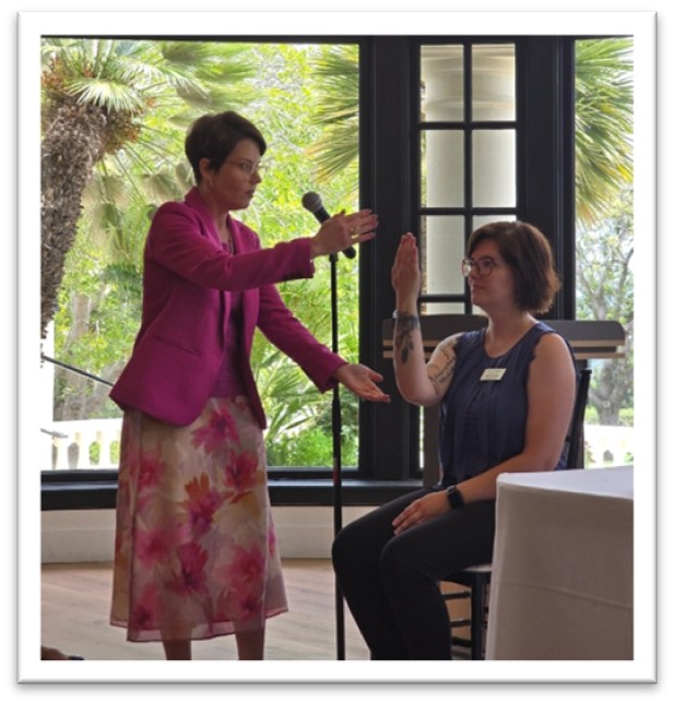 A woman in a pink blazer gestures toward a seated woman holding her hand upright in front of a microphone, inside a sunlit room with large windows and palm trees outside.