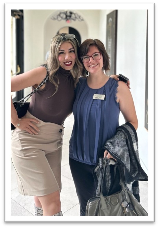 Two women stand together indoors, smiling at the camera. One wears a brown top and tan skirt, the other wears glasses, a blue blouse, and a name tag. Both appear friendly and happy.