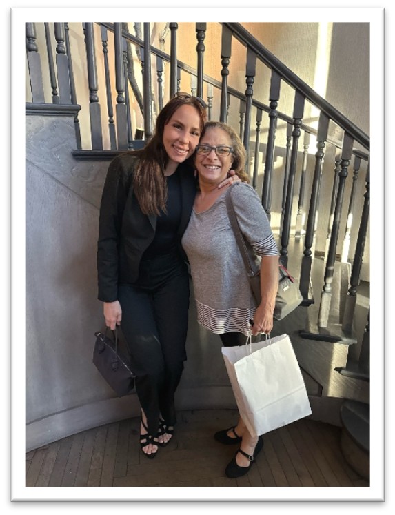Two women smile and pose together by a staircase indoors. One woman wears black and holds a purse; the other wears a gray top, glasses, and holds a white shopping bag.