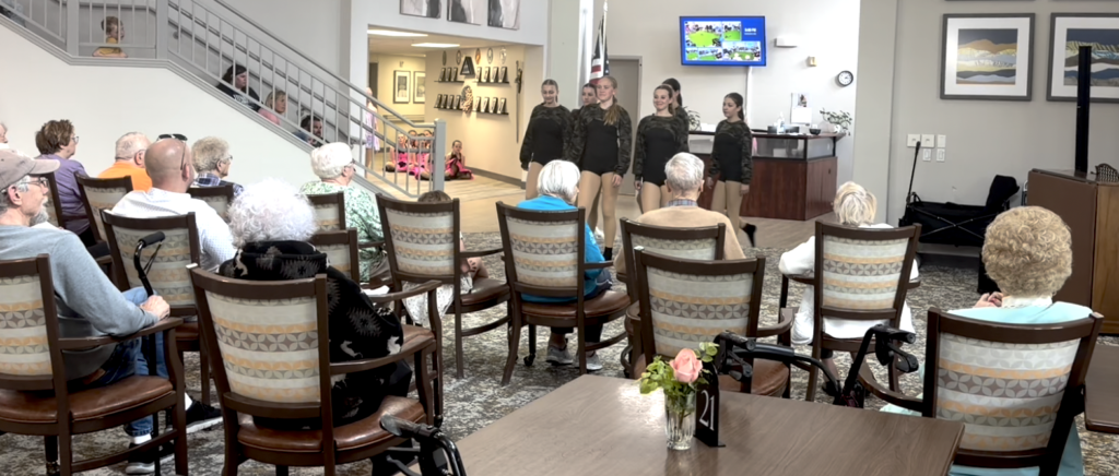 A group of young dancers performs for an audience of seniors seated in chairs in a common room. The room has a staircase, artwork on the walls, a TV, and floral arrangements on the tables.