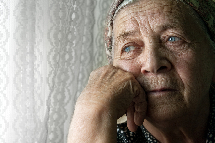 An elderly woman with blue eyes and a headscarf rests her chin on her hand, gazing thoughtfully out a window with sheer lace curtains.