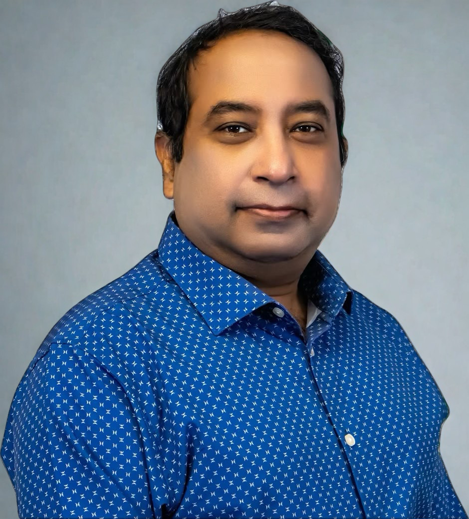 A man with short black hair wearing a blue button-up shirt with a white pattern, posing in front of a plain light gray background.