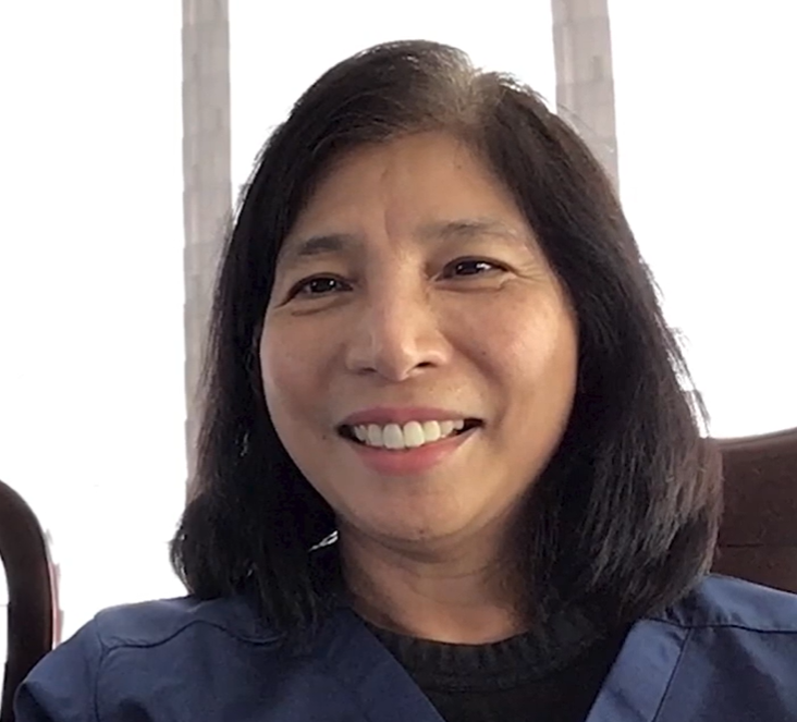 A woman with straight, shoulder-length black hair smiles at the camera while wearing a blue top, seated indoors with bright light coming from behind.