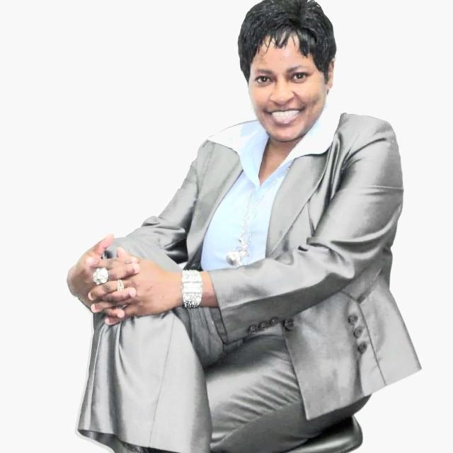 A woman in a gray suit and light blue shirt sits on a stool with her legs crossed, smiling at the camera. She has short hair and wears jewelry, including rings, a watch, and a necklace.