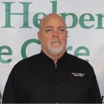A man with a bald head and gray beard wearing a black collared shirt stands in front of a banner with partially visible green text.