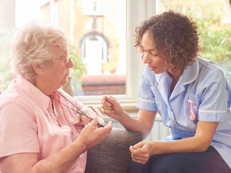 A nurse in a blue uniform speaks kindly to an older woman with gray hair and glasses, who is sitting on a couch and wearing a pink shirt. Sunlight streams through large windows in the background.