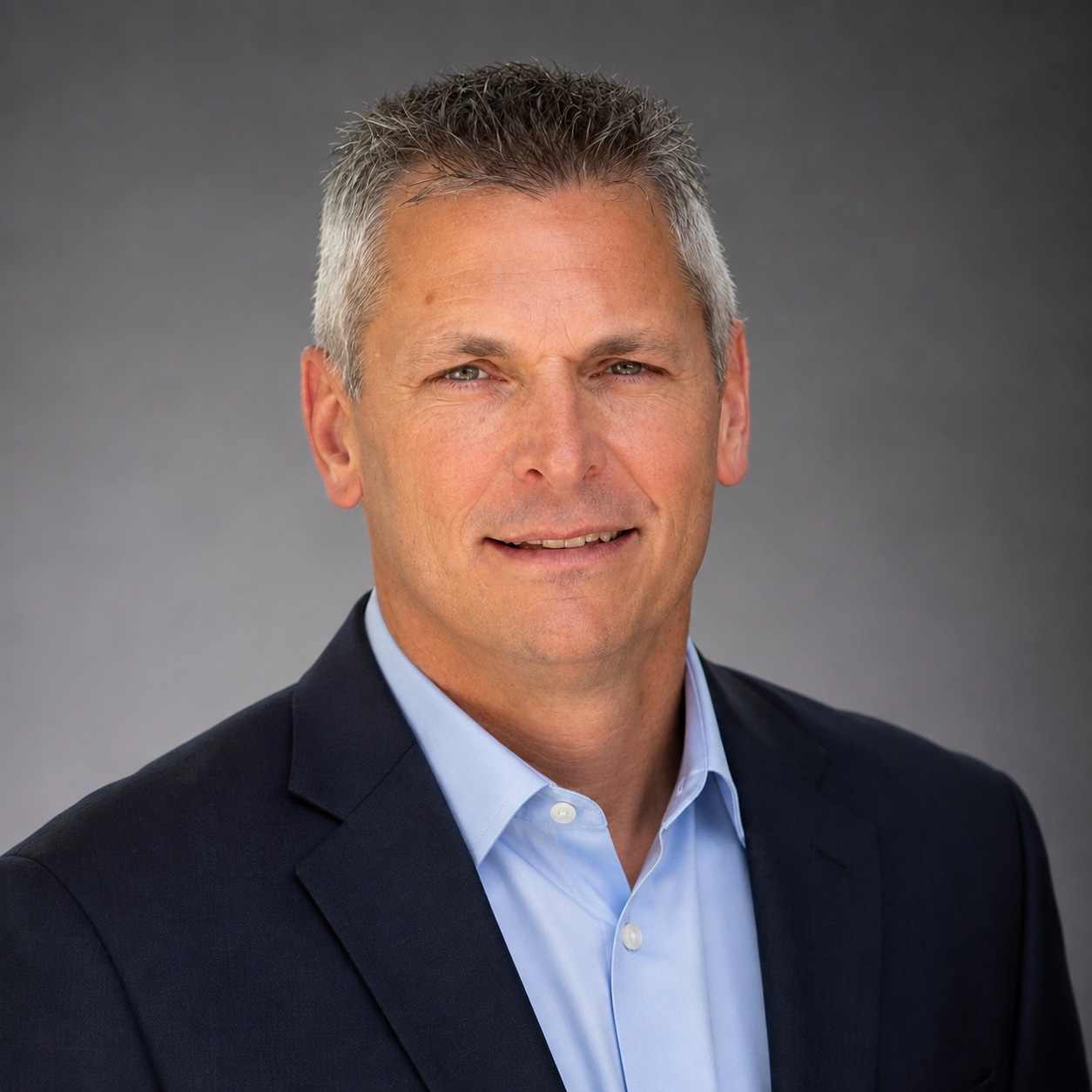 A middle-aged man with short gray hair wearing a navy suit jacket and light blue shirt, smiles slightly while posing in front of a plain gray background.