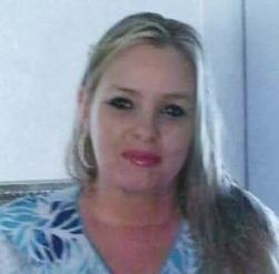 A woman with long blonde hair and hoop earrings, wearing a light blue floral top, is standing indoors against a plain light-colored background.