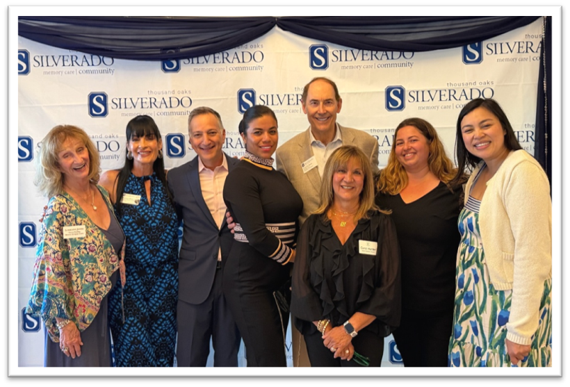 Eight people stand together smiling in front of a Silverado Memory Care event backdrop, dressed in business or semi-formal attire, posing for a group photo.