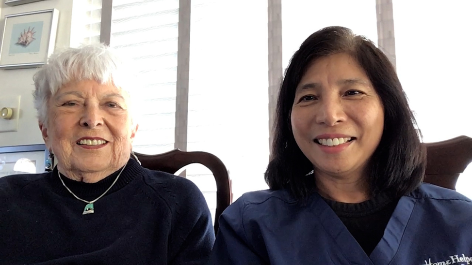 Two women smiling while sitting indoors. One has short white hair and a dark sweater; the other has straight black hair and wears dark blue scrubs. Bright window blinds and framed pictures are visible in the background.