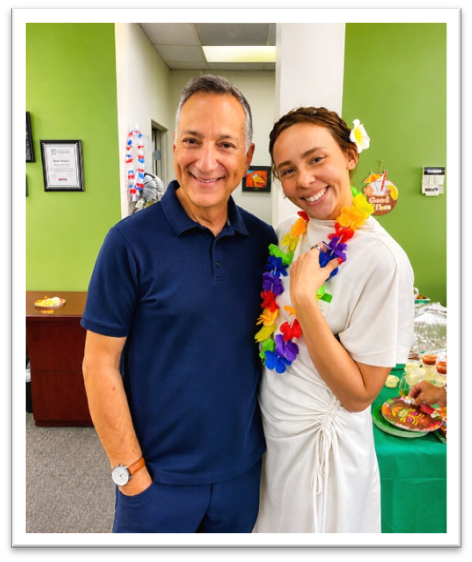 Two people stand smiling in an office. The man wears a navy shirt and pants; the woman wears a white dress, a colorful flower lei, and a flower in her hair. A table with food and green decor is behind them.