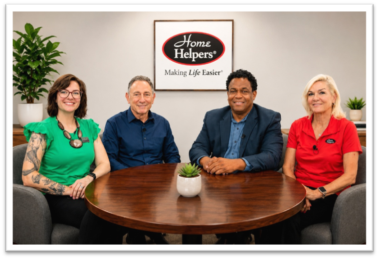 Four people sit at a round table in front of a sign that reads Home Helpers Making Life Easier. They are smiling, with plants in the background and a small plant on the table.