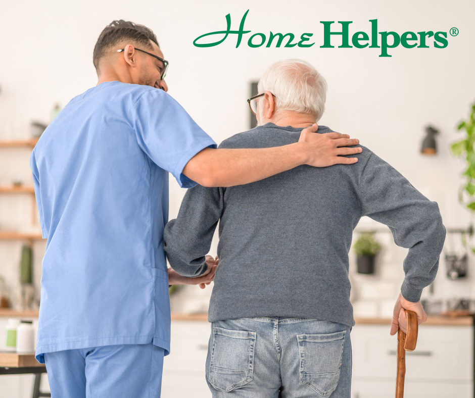 A caregiver in blue scrubs supports an elderly man with a cane. The Home Helpers logo appears at the top. They are indoors in a bright, comfortable setting.