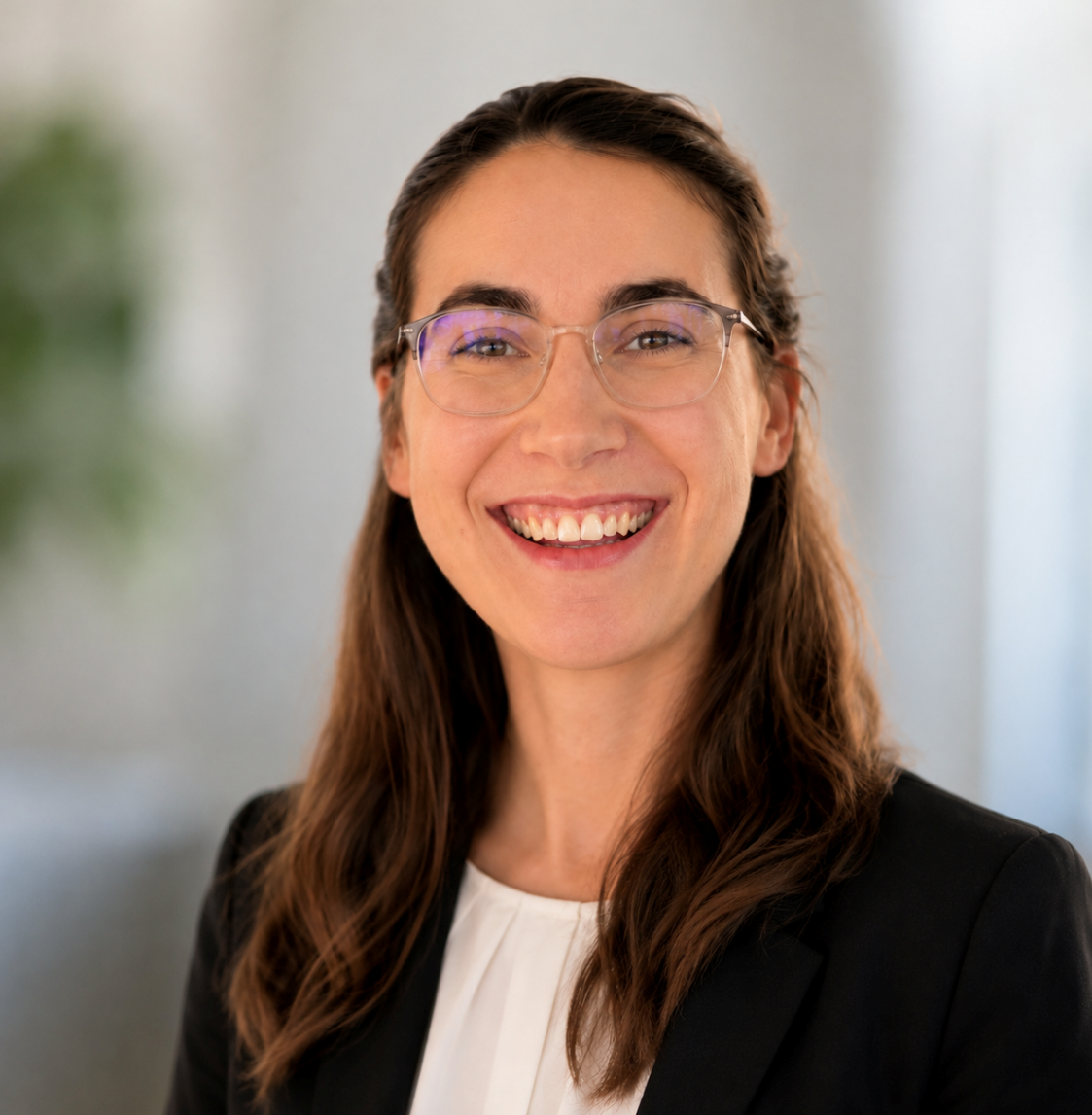 A woman with long brown hair and glasses smiles while wearing a black blazer and white blouse. The background is softly blurred.