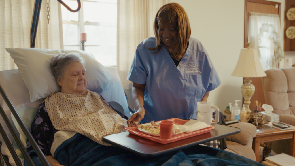 A nurse in blue scrubs assists an elderly woman in a hospital bed, serving her a meal on a tray. The room appears homey, with sunlight coming through curtains and comfortable furniture nearby.
