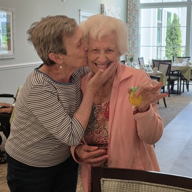 Two older women are smiling; one kisses the other on the cheek while she holds a decorated cookie. They seem happy in a bright, cheerful dining area.