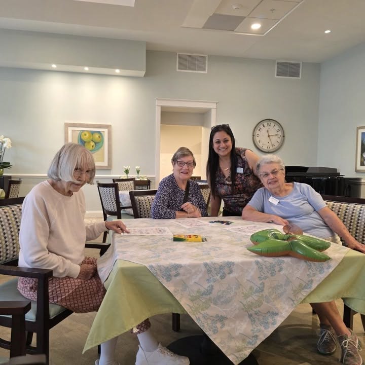 Three elderly women and a younger woman sit and smile around a table in a bright room. Art supplies and a puzzle are on the table. A piano and a clock are visible in the background.