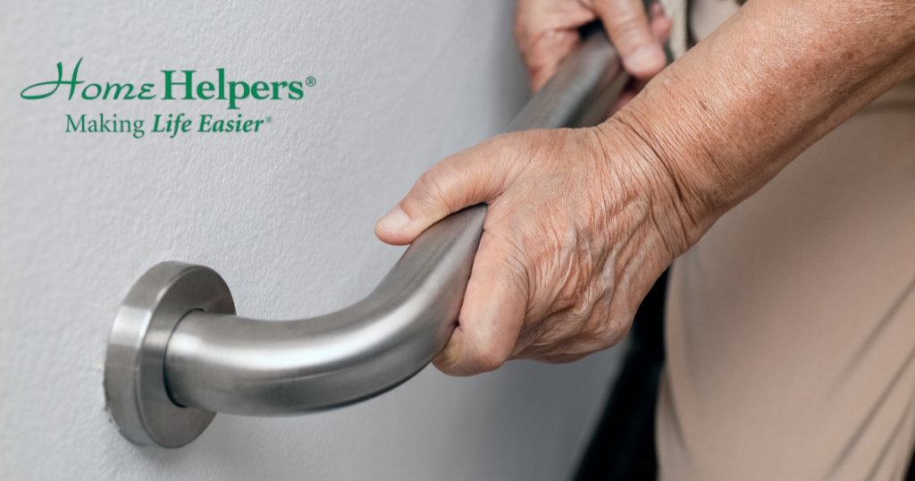 Close-up of an elderly persons hands holding onto a stainless steel grab bar, with the Home Helpers Making Life Easier logo in green text on the wall in the background.