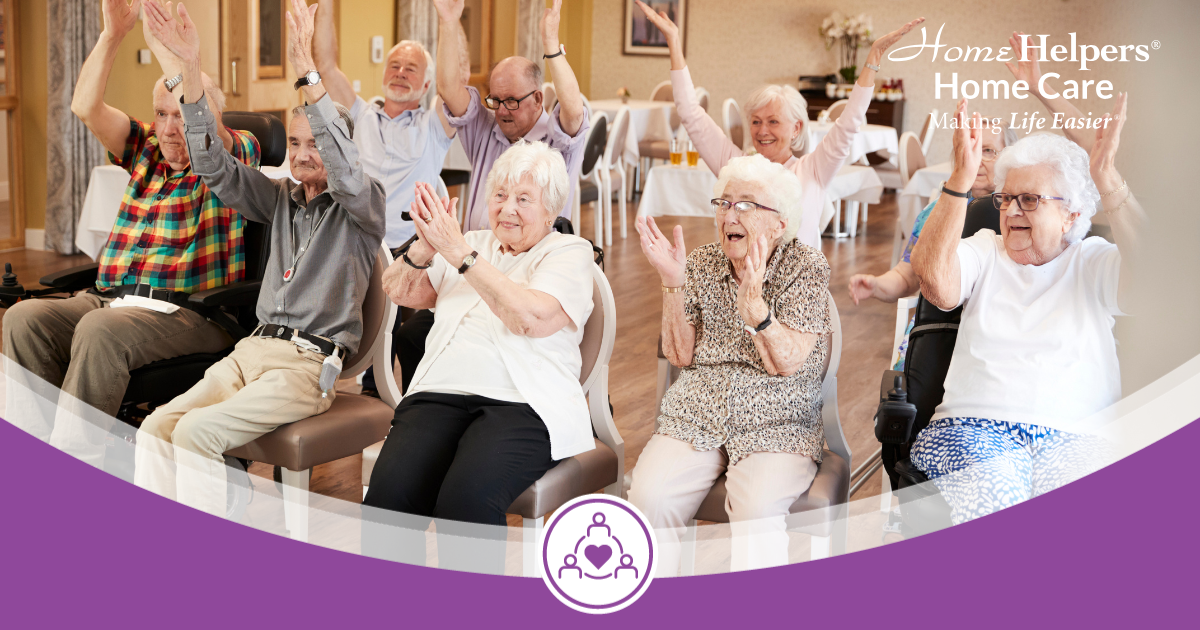 A group of smiling elderly people sit in chairs with arms raised, participating in a joyful group activity at a care facility. Home Helpers Home Care logo appears in the top right corner.