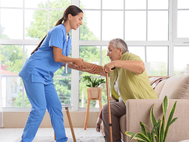 A nurse in blue scrubs helps an elderly woman with a cane stand up from a sofa in a bright living room with large windows and plants.